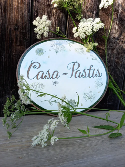 Vollständiges ovales Schild „Casa Pastis“ vor dunklem Holz, daneben weiße Blüten.